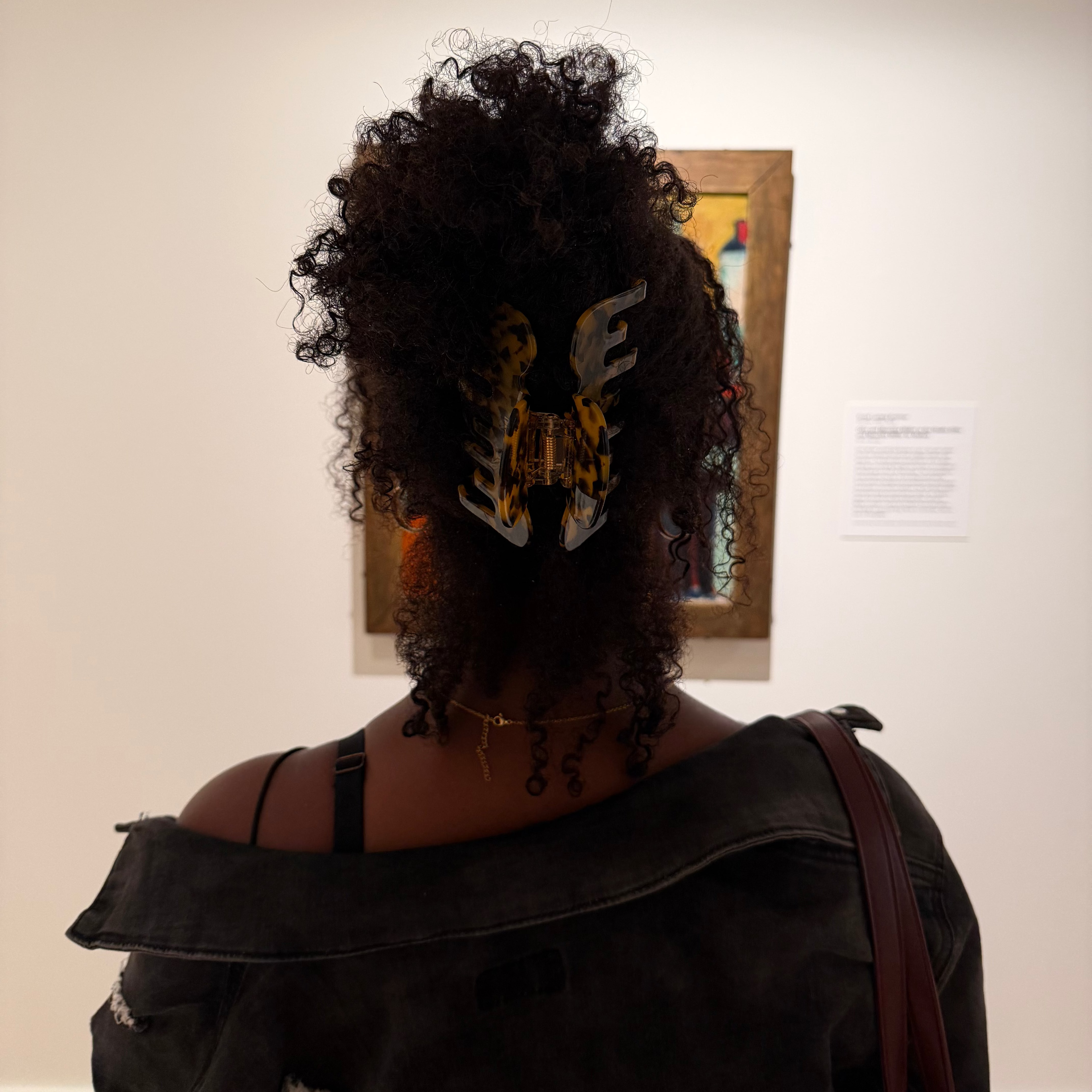 A person viewed from behind in a gallery or museum setting, wearing a large black denim jacket and a black top, with a voluminous curly black wig styled with a decorative claw clip. A framed artwork hangs on the white wall behind them.