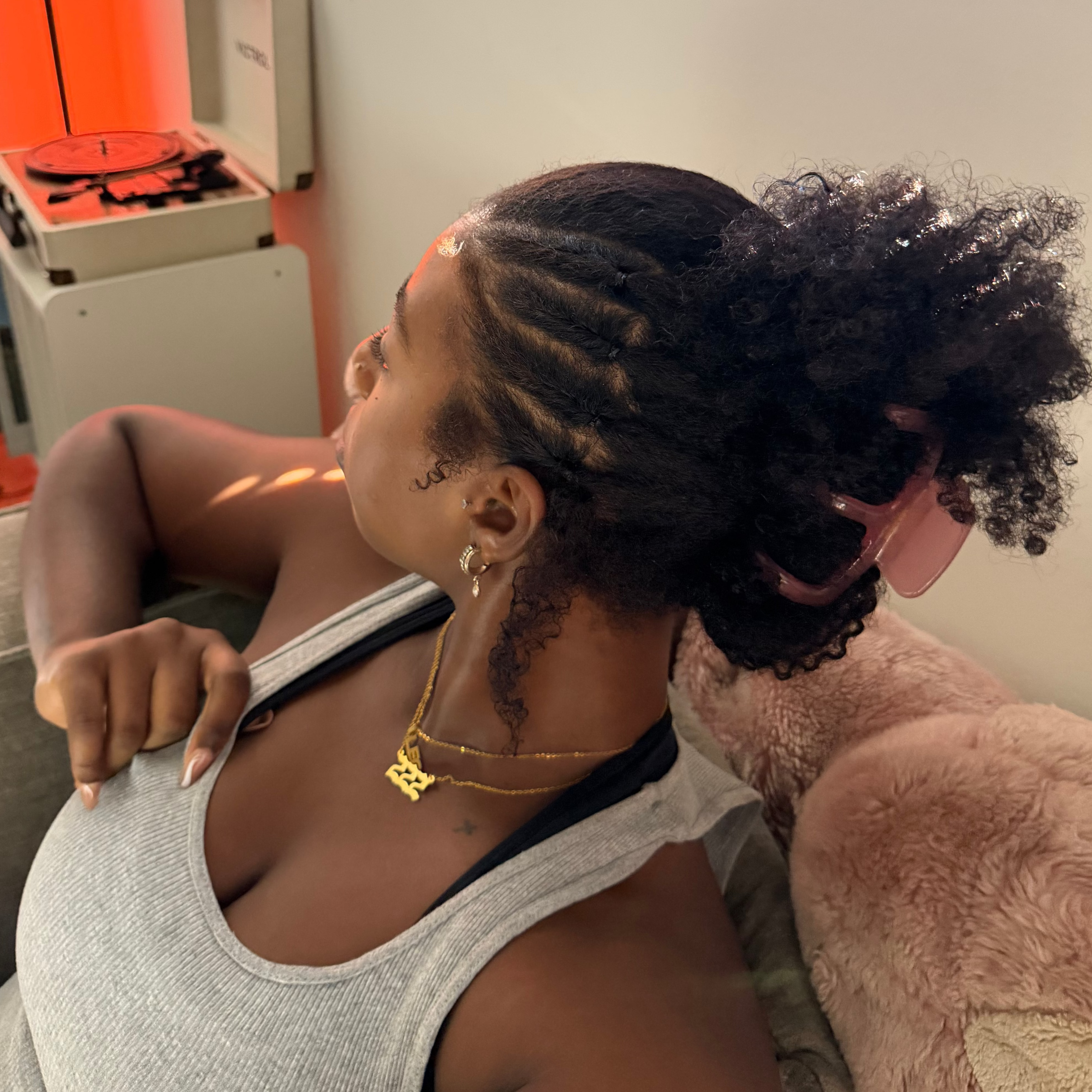 A woman with styled black cornrow hair and a high puff bun secured with a pink claw clip, wearing a gray tank top and gold necklaces, sitting indoors next to a pink plush toy.