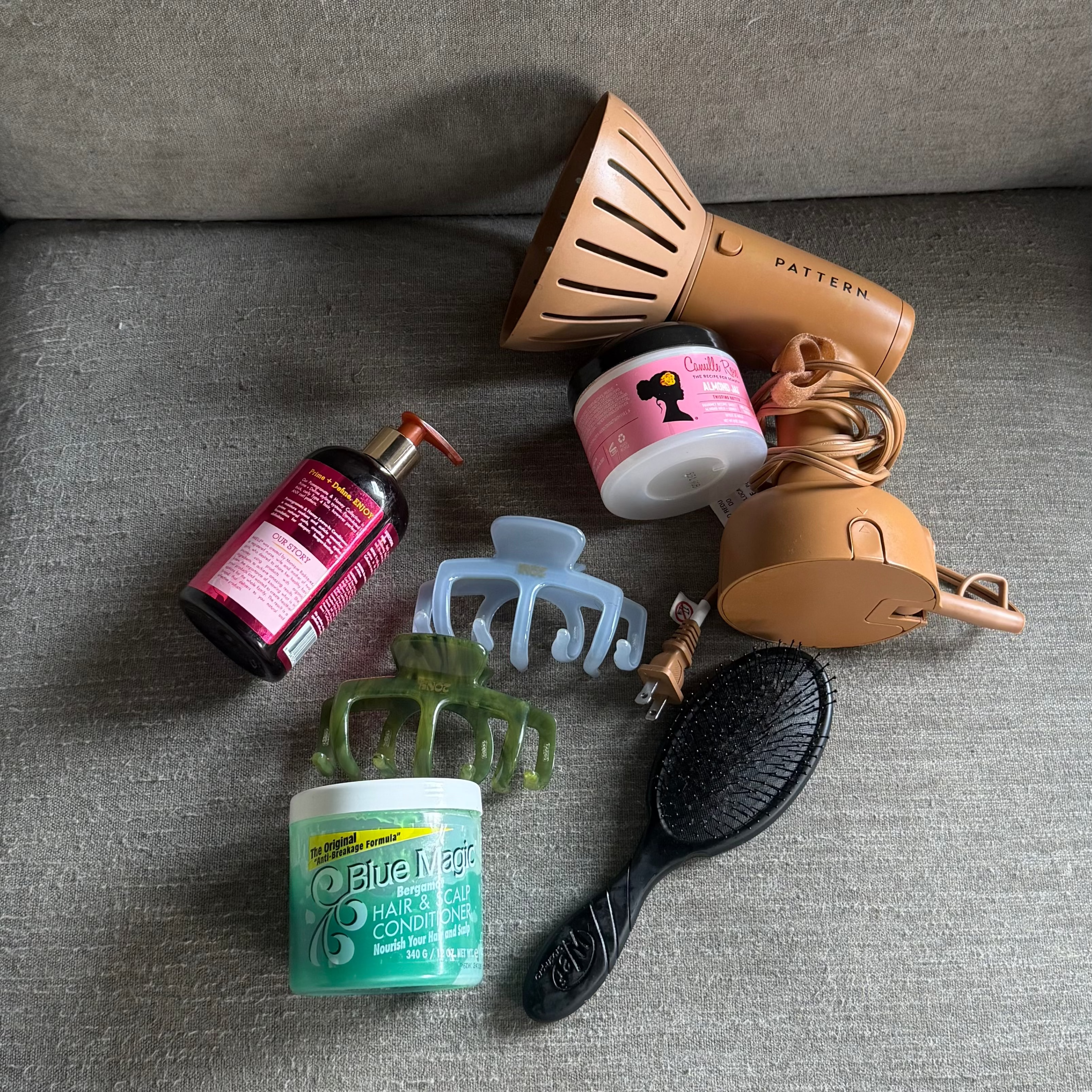 A collection of hair care and styling products arranged on a gray fabric sofa, including a pink bottle, a brown hair dryer, a pink and white hair marker, a green claw clip, a light blue claw clip, a green hair scrunchie, a white hair product jar, and a black hairbrush.